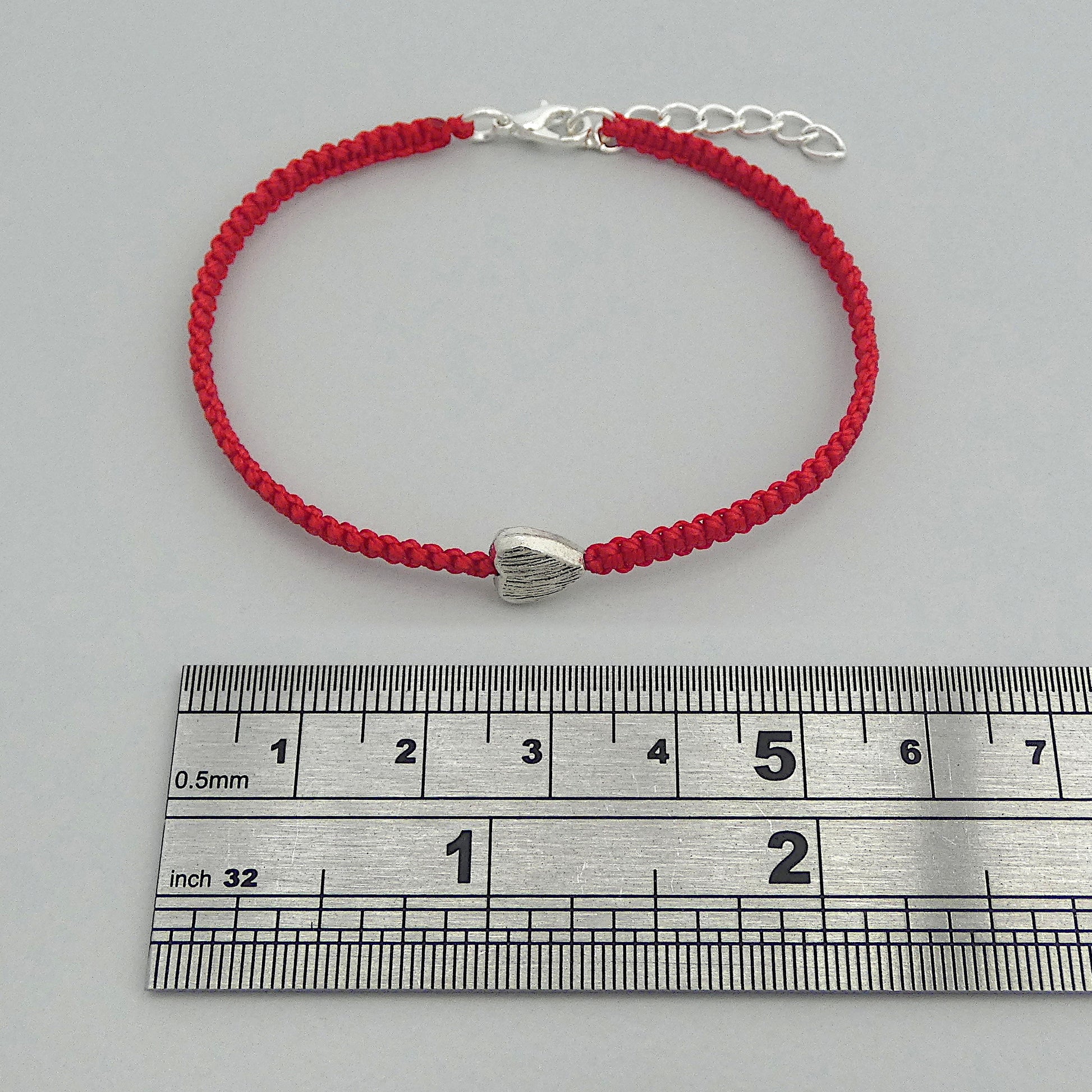 Red seven inch macramé bracelet with silver plated textured heart charm, lobster clasp and one inch extender chain next to a ruler.