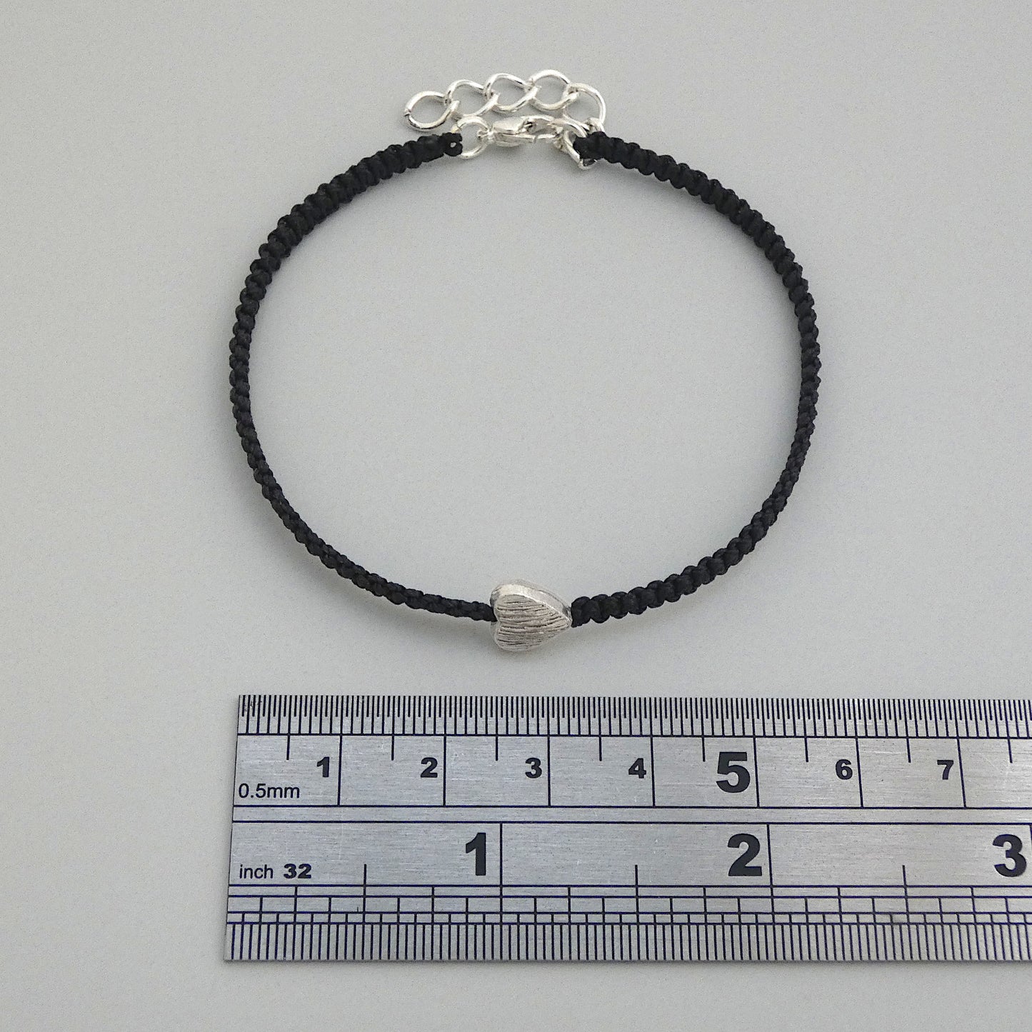 Black seven inch macramé bracelet with silver plated textured heart charm, lobster clasp and one inch extender chain next to a ruler.