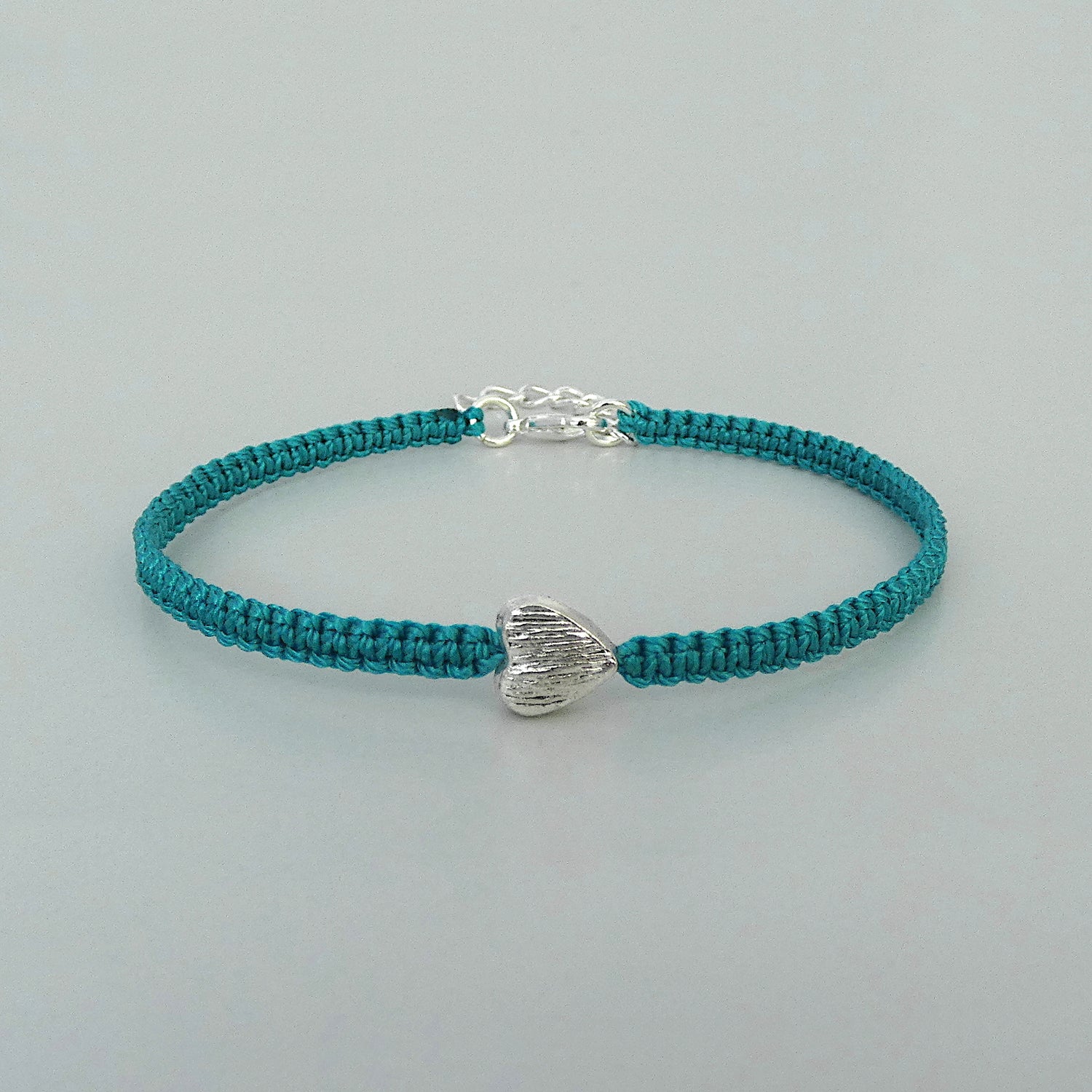 Teal green seven inch macramé bracelet with silver plated textured heart charm, lobster clasp and one inch extender chain.